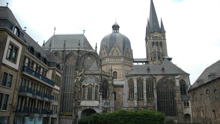 Eine beeindruckende Kirche mit markanten Türmen und gotischer Architektur. Die Gebäude umgeben einen kleinen Platz mit einigen modernen Wohnhäusern.