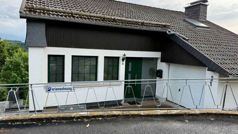 Ein modernes Ferienhaus mit weißen Wänden und einem schrägen Dach. Die Fassade zeigt große Fenster und ein Schild mit der Aufschrift „Ferienwohnung 2“.