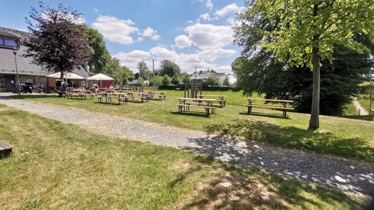 Ein grüner Garten mit mehreren Tischen und Bänken. Der Himmel ist blau mit vereinzelten Wolken.