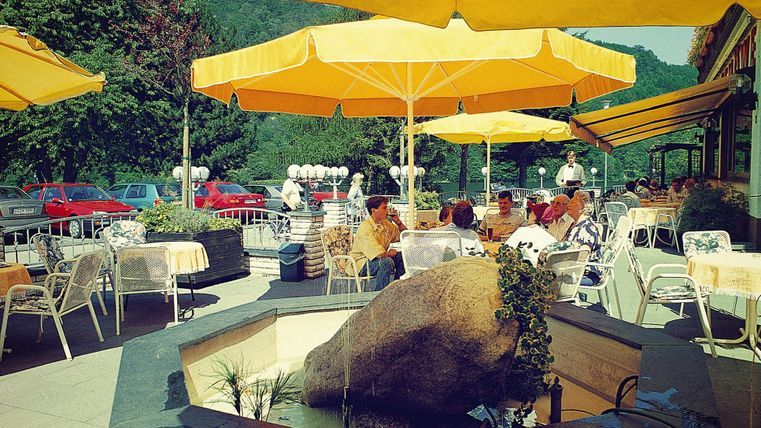 Ein Cafe im Freien mit gelben Sonnenschirmen und einem kleinen Brunnen. Gäste sitzen an Tischen und genießen die Umgebung.