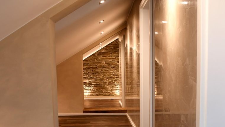 Ein moderner Flur mit schrägen Wänden und angenehmer Beleuchtung. Der Boden ist aus Holz und es gibt eine Steinwand im Hintergrund.