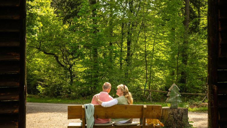 Ein Paar sitzt auf einer Bank im Wald und genießt die Natur. Die Umgebung ist grün und sonnig, ideal für einen entspannten Moment.