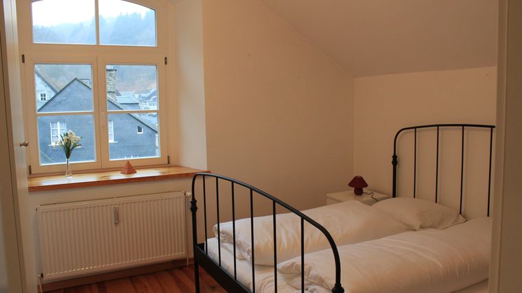 Ein einfaches Schlafzimmer mit einem Metallbett und frisch bezogenen Bettdecken. Das Fenster bietet Blick auf eine ruhige Landschaft und einen kleinen Blumenstrauß steht auf dem Fensterbrett.