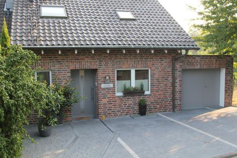 Ein modernes, zweigeschossiges Haus aus Backstein mit einem Satteldach. Vor dem Haus gibt es einen gepflasterten Weg und einige Pflanzen in Töpfen.