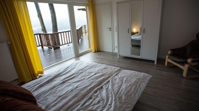 Ein heller Raum mit einem Bett und großen Fenstern, die auf eine Terrasse blicken. Gelbe Vorhänge verleihen dem Raum eine freundliche Atmosphäre.