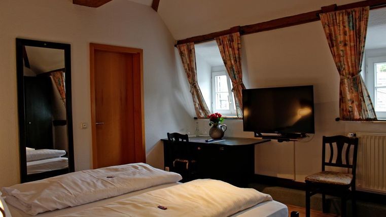 Ein gemütliches Zimmer mit zwei Betten, einem Spiegel und einem Fernseher. Große Fenster mit Vorhängen und ein einfacher Tisch vervollständigen die Einrichtung.