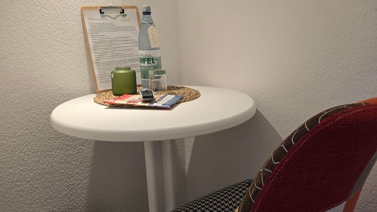 Ein kleiner Tisch mit einer bunten Stuhl, Flasche Wasser und Notizblock. Neben dem Tisch steht ein Mülleimer.