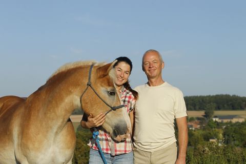 Eine Frau und ein Mann stehen nebeneinander und lächeln. Neben ihnen befindet sich ein Pferd vor einer malerischen Landschaft.