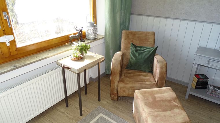 Ein gemütlicher Raum mit einem bequemen Sessel und einem kleinen Tisch. An einem Fenster mit natürlichem Licht stehen Pflanzen und eine dekorative Lampe.