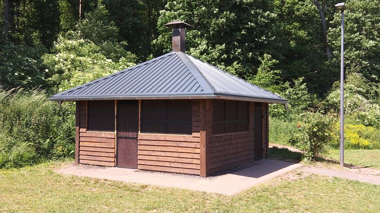 Ein Holzhaus mit einem grauen Dach steht in einer grünen Umgebung. Umgeben von Bäumen und Sträuchern bietet es einen gemütlichen Rückzugsort.