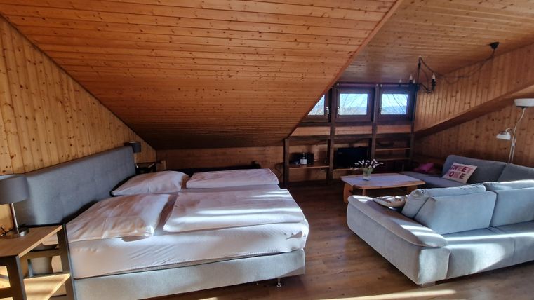 Ein gemütlicher Dachraum mit zwei Betten und einem Sofa. Die Holzverkleidung und großen Fenster schaffen eine einladende Atmosphäre.