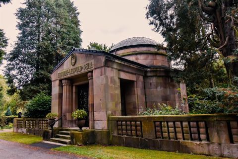 Ein historisches Mausoleum aus Stein, umgeben von Bäumen und grünem Gras. Die Architektur zeigt klare Linien und einen runden Dachaufsatz.
