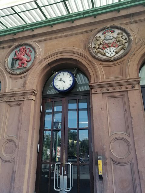 Ein historisches Gebäude mit einem großen Eingang und einer runden Uhr. Über dem Eingang sind zwei Wappen zu sehen.