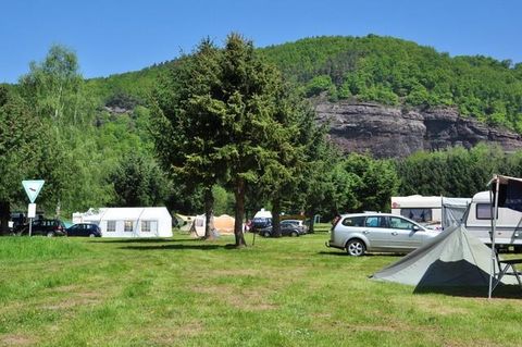 Ein Campingplatz mit grünen Wiesen, Bäumen und Wohnwagen. Im Hintergrund erhebt sich eine bewaldete Berglandschaft unter blauem Himmel.