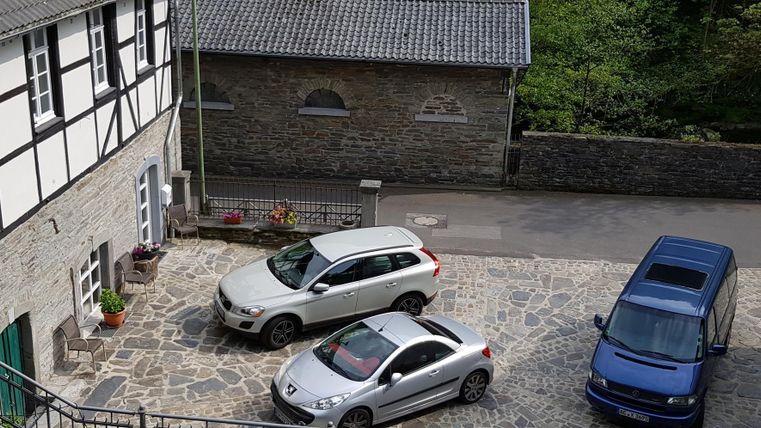 Ein gepflasterter Innenhof mit drei Autos und einem historischen Gebäude im Hintergrund. Die Umgebung ist grün und ruhig.