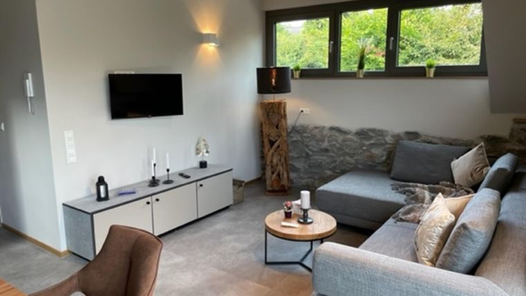 Ein modernes Wohnzimmer mit einem grauen Sofa und einem Tisch. Große Fenster sorgen für viel Tageslicht und einen Blick ins Grüne.