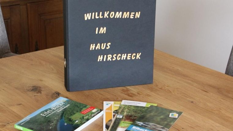 Ein schwarzes Ordner mit der Aufschrift "Willkommen im Haus Hirschcheck" liegt auf einem Holztisch. Daneben befinden sich einige Broschüren über die Umgebung.