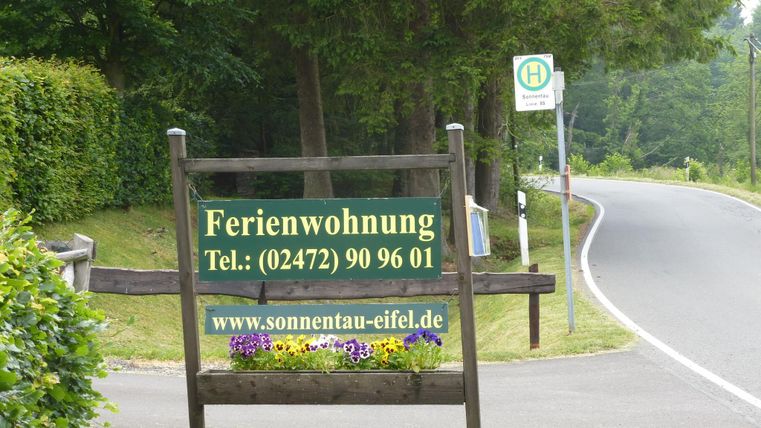 Ein Schild für eine Ferienwohnung steht an einer ruhigen Straße. Im Hintergrund sind Bäume und eine Bushaltestelle sichtbar.