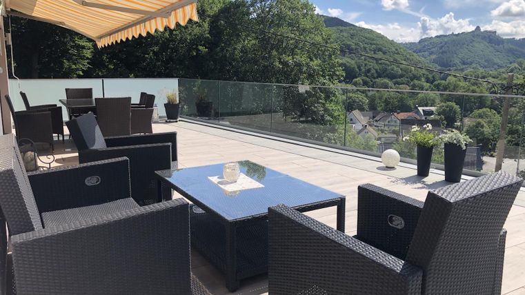 Eine moderne Terrasse mit einem schwarzen Rattan-Sitzbereich und einem Glastisch. Im Hintergrund sind grüne Hügel und strahlend blauer Himmel zu sehen.