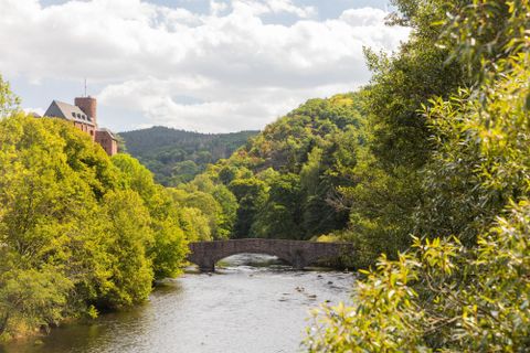 Eine malerische Landschaft mit einem Fluss und einer Bogenbrücke. Umgeben von grünen Bäumen und sanften Hügeln unter einem bewölkten Himmel.