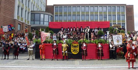Eine festliche Parade mit Kostümierten und Fahnen. Im Hintergrund eine bunte Bühne mit vielen Teilnehmern und einer festlichen Atmosphäre.