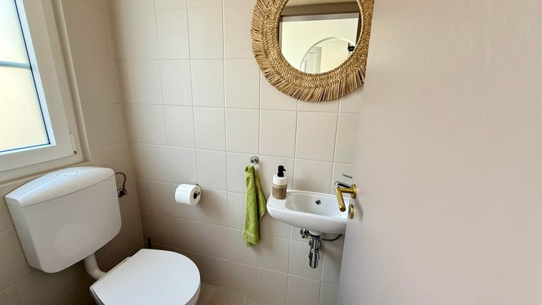 Ein kleines, helles Badezimmer mit einer Toilette und einem Waschbecken. Ein runder Spiegel aus natürlichem Material hängt an der Wand.