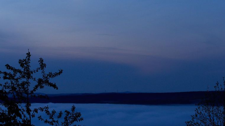 Ein geheimnisvoller Nachthimmel mit einer teilweise verdeckten Mondscheibe. Darunter breitet sich eine dichte Nebelschicht über die Landschaft aus.