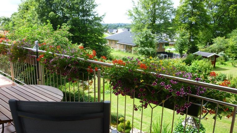 Ein schöner Balkon mit blühenden Pflanzen und Blick ins Grüne. Im Hintergrund sind Einfamilienhäuser und Bäume sichtbar.