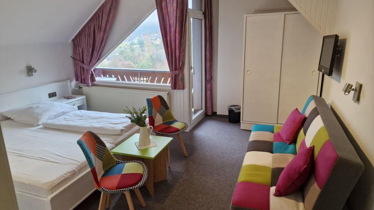 Ein helles Zimmer mit einem Fenster und schöner Aussicht. Es gibt ein Bett, einen Tisch mit bunten Stühlen und ein Sofa.
