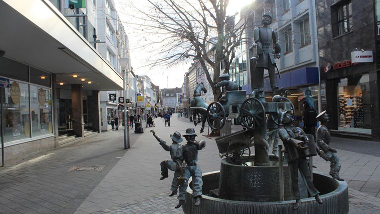 Eine lebendige Einkaufsstraße mit einem Brunnen in der Mitte. Die Statue zeigt fröhliche Figuren und umgeben von Geschäften und Passanten.