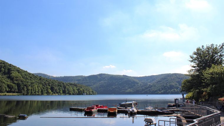 Ein ruhiger See umgeben von grünen Hügeln. Am Ufer sind Boote und eine schöne Promenade zu sehen.