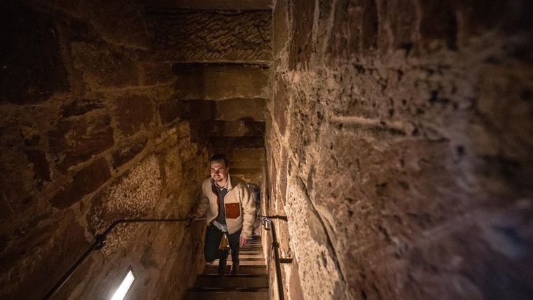 Eine Person steigt eine steinerne Treppe hinauf. Die Wände sind aus grobem Stein und das Licht ist gedämpft.