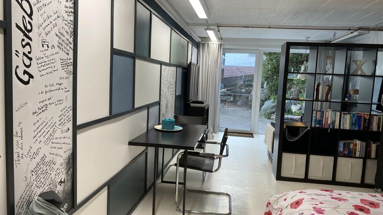 Ein modernes Zimmer mit einem Tisch und Stühlen sowie Regalen für Bücher. Große Fenster sorgen für viel Tageslicht.