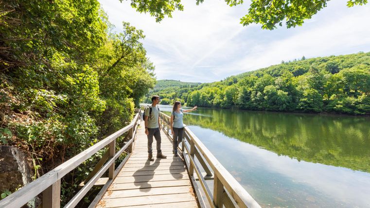 Ein Wanderweg an einem ruhigen Fluss umgeben von üppigem Grün. Zwei Personen erkunden die Natur und genießen die Aussicht.