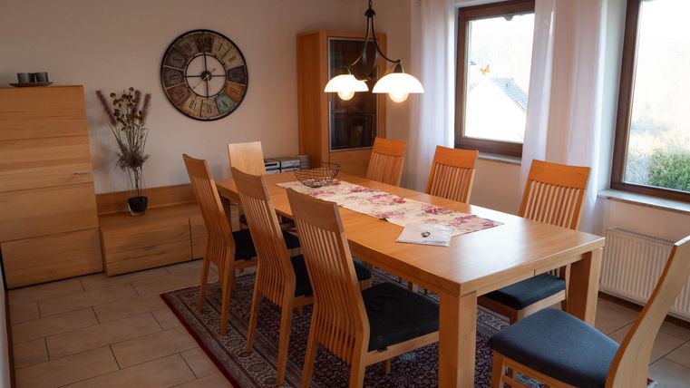 Ein heller Essbereich mit einem großen Holztisch und zehn Stühlen. An den Wänden hängen Fenster und ein runder Wandkalender.