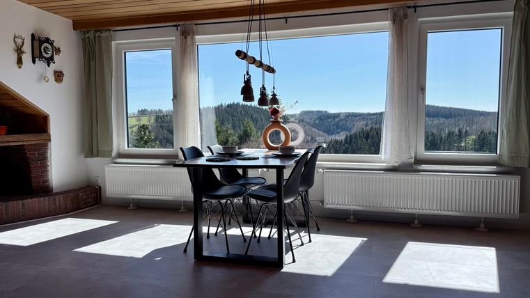 Ein modernes Esszimmer mit einem Tisch und Stühlen, das von großen Fenstern umgeben ist. Durch die Fenster hat man einen schönen Blick auf die bewaldete Landschaft.