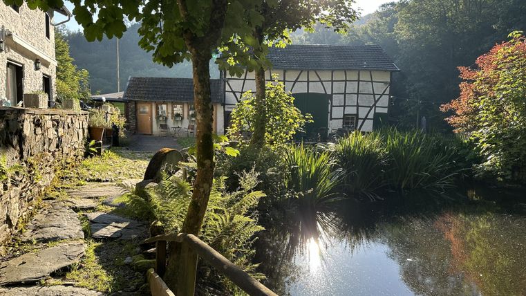 Ein malerischer Teich umgeben von üppigem Grün und Bäumen. Im Hintergrund ist ein traditionelles Haus mit Fachwerkstil zu sehen.