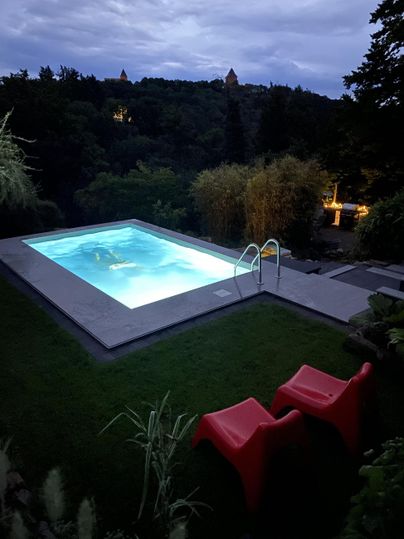 Ein moderner Pool in einem grünen Garten unter einem Abendhimmel. Rote Stühle laden zum Entspannen ein.