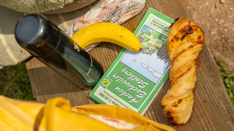 Ein Picknick mit einer Brotrolle, einer Banane und einer Thermoskanne auf einem Holztisch. Die Umgebung scheint sonnig und entspannend zu sein.