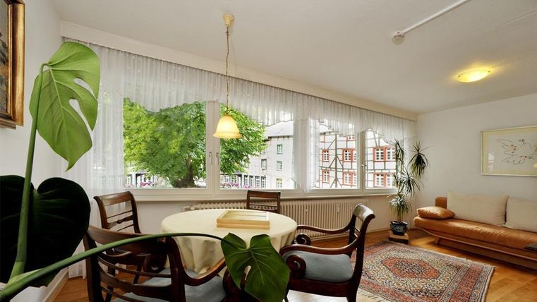 Ein heller und einladender Wohnraum mit einem runden Tisch und Stühlen. Große Fenster bieten einen Blick auf Bäume und ein ruhiges Umfeld.