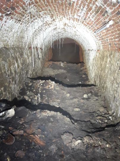 Ein dunkler Tunnel mit roten Ziegelwänden und einem unebenen Boden. Einige Latten und Schmutz sind auf dem Boden verteilt.