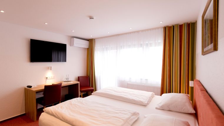 Ein modernes Hotelzimmer mit einem Doppelbett und einem Fernseher an der Wand. Der Raum hat große Fenster mit farbigen Vorhängen und einen kleinen Arbeitsbereich.
