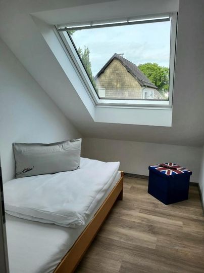 Ein gemütliches Zimmer mit einem Einzelbett und einem Fenster, das Tageslicht hereinlässt. Neben dem Bett steht eine Sitzgelegenheit mit einem britischen Motiv.