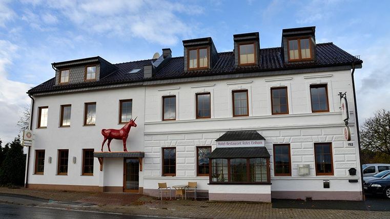 Ein modernes Restaurantgebäude mit weißer Fassade und einem roten Reh als Skulptur an der Vorderseite. Die Fenster sind ausgestattet mit braunen Rahmen und das Gebäude hat ein schickes, einladendes Aussehen.