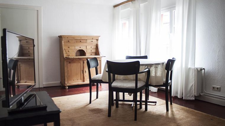 Ein heller Raum mit einem Tisch und Stühlen. Im Hintergrund steht eine elegante Kommode und große Fenster lassen viel Licht herein.