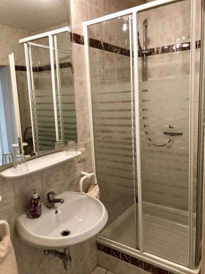Ein modernes Badezimmer mit einer Glastusche und einem Waschbecken. Die Wände sind hell gefliest und es gibt einen Spiegel.