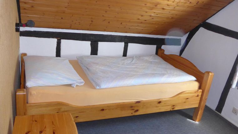 Ein einfaches Schlafzimmer mit einem Holzbett und beige Bettwäsche. Die Wände sind teilweise mit Holzverkleidung und die Decke ist schräg.