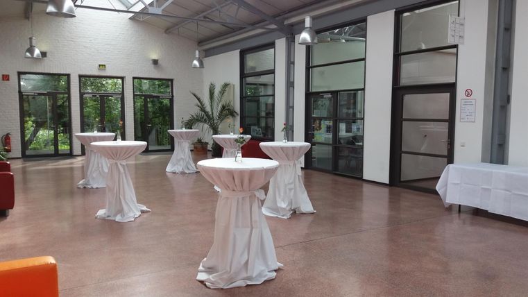 Ein heller Veranstaltungsraum mit hohen Fenstern und modernen Stehtischen. Die Szene ist einladend und gut geeignet für Events oder soziale Zusammenkünfte.