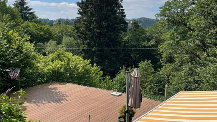 Eine ruhige Terrasse umgeben von üppigem Grün und Bäumen. Im Hintergrund sind sanfte Hügel zu sehen, und es gibt eine Basketballkorbanlage.