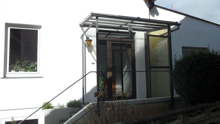 Ein modernes Eingangsportal mit einer Überdachung aus Glas. Die Fassade des Hauses ist weiß und hat große Fenster.